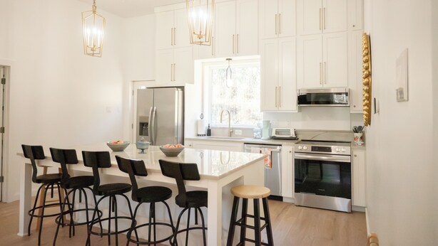 Una cocina moderna con armarios blancos, una isla central con taburetes, electrodomésticos de acero inoxidable e iluminación colgante.