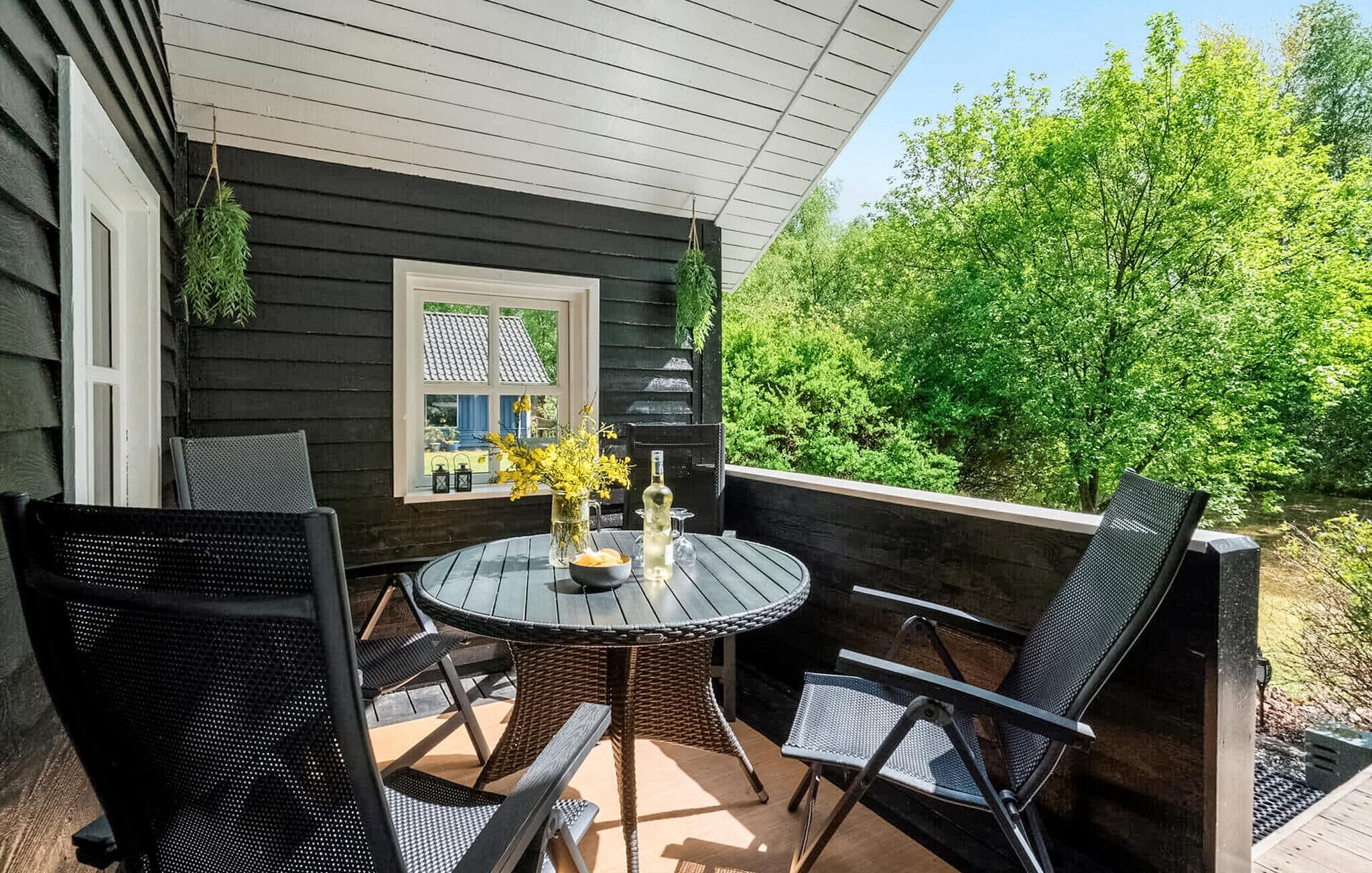 Un patio techado con una mesa redonda y cuatro sillas, una ventana con vista a la vegetación y plantas colgantes.