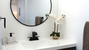 Une salle de bain avec un miroir rond, un lavabo, un porte-serviettes et une petite plante orchidée.