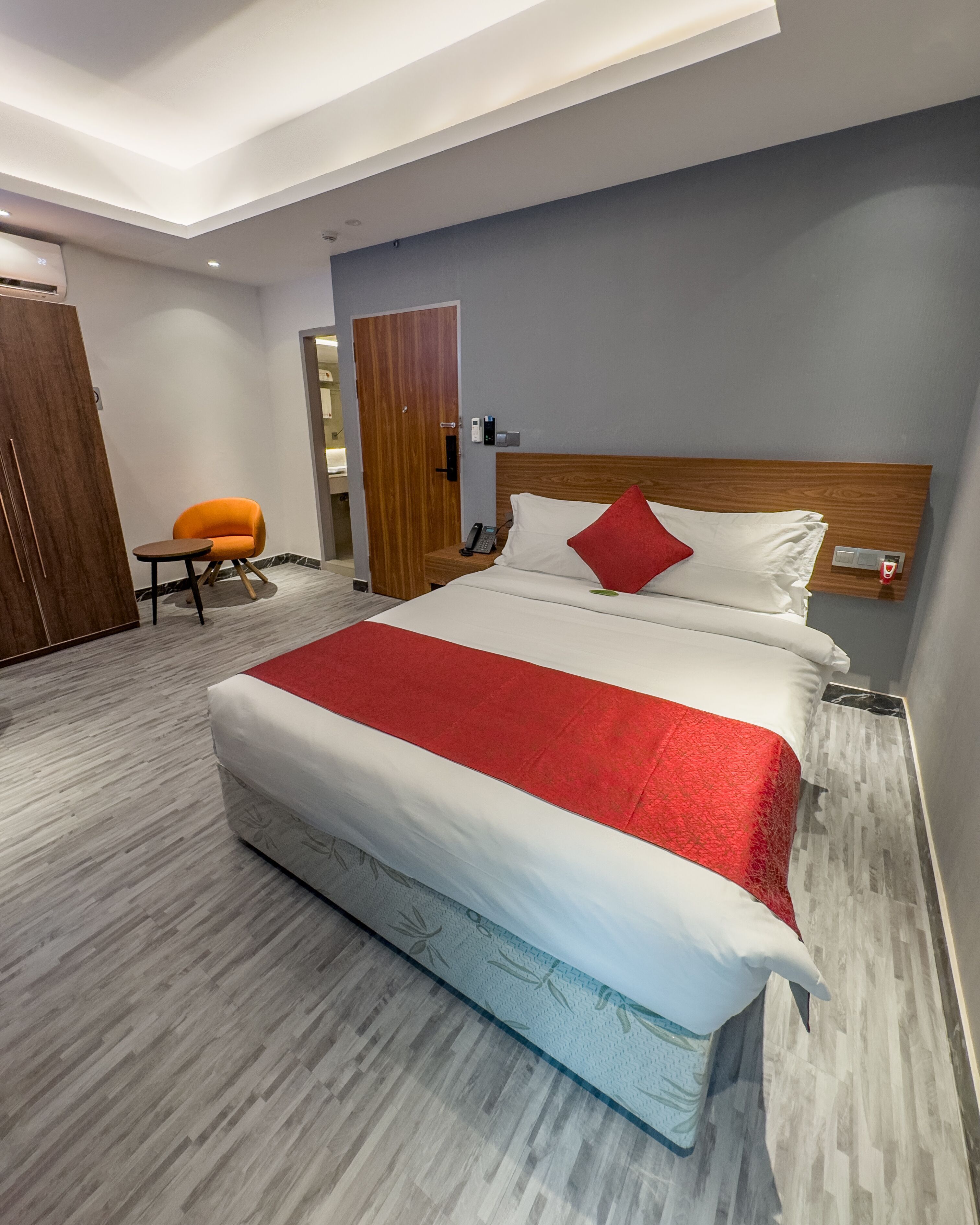 A modern hotel room with a large bed, a wooden headboard, a small table with a chair, and a red accent wall.
