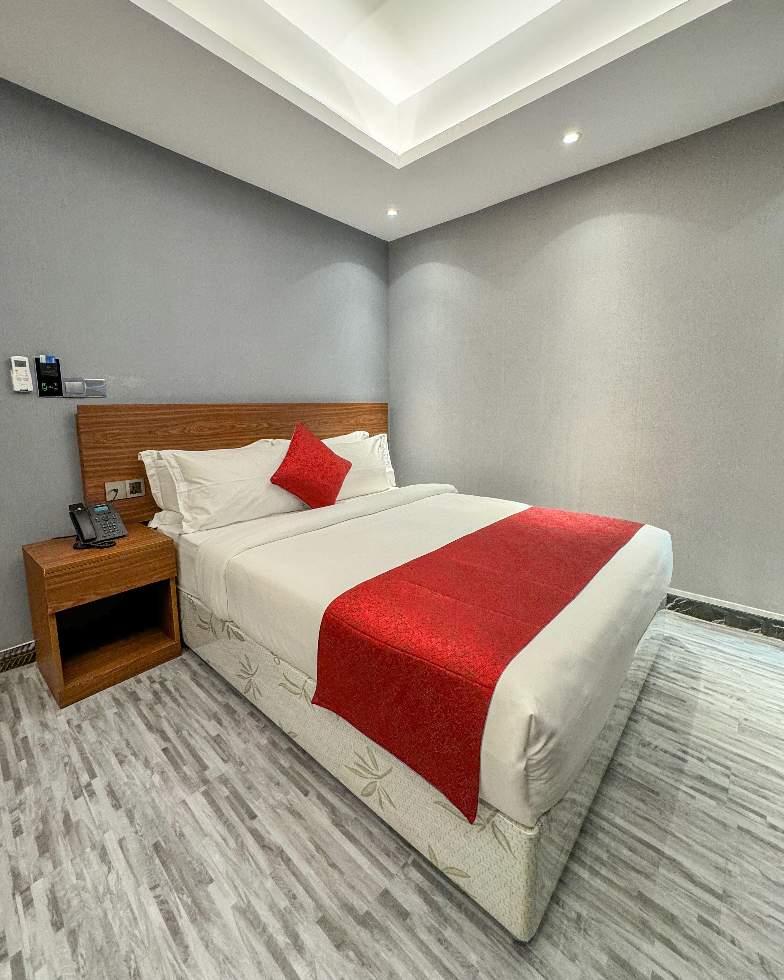 A neatly made bed with a red throw, a wooden nightstand with a phone, and a wall-mounted control panel.