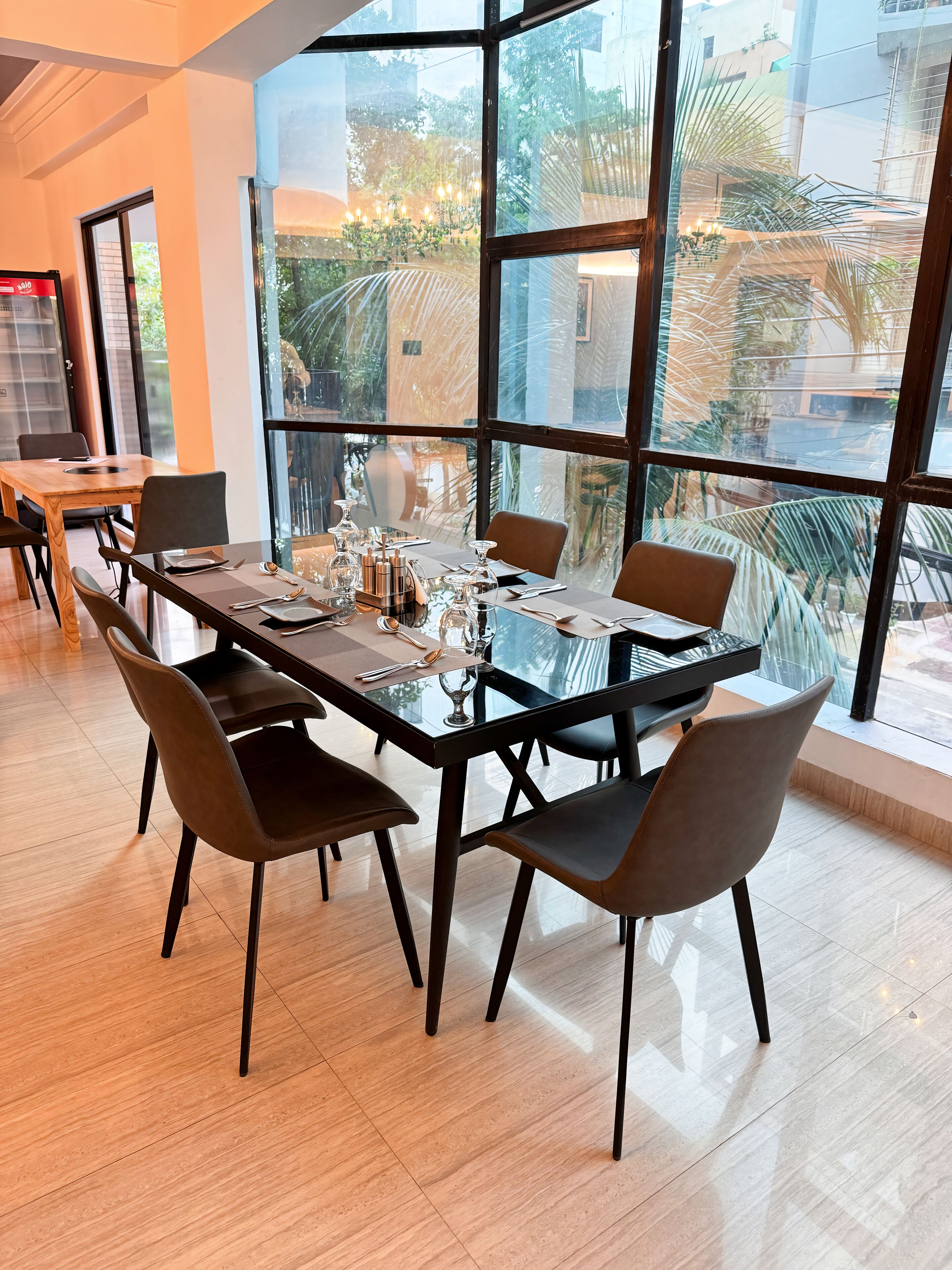 A modern dining area with a glass table and chairs, large windows offering a view of greenery and buildings outside.
