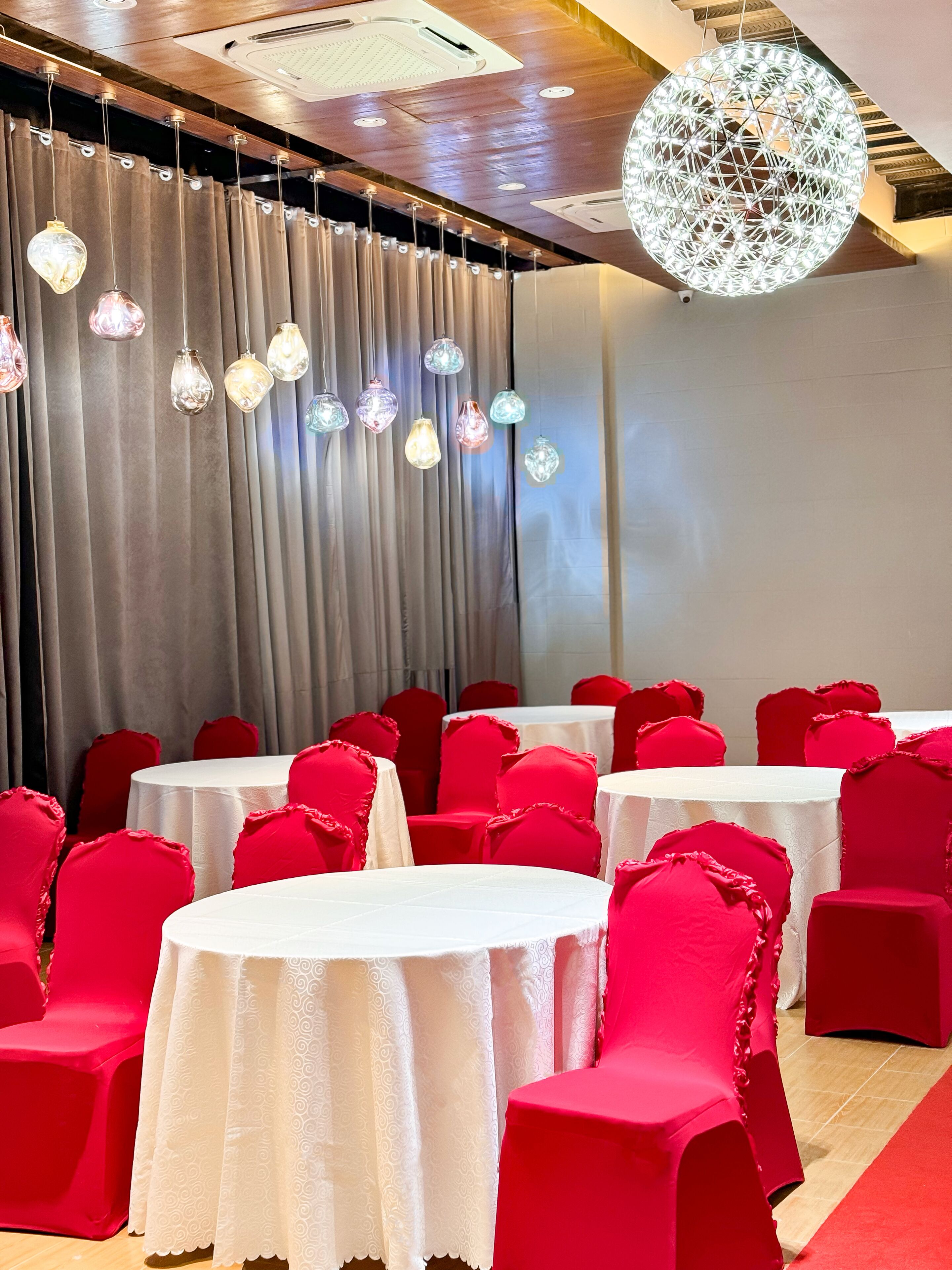 A banquet hall with round tables covered in white tablecloths, red chairs, and a decorative ceiling light.
