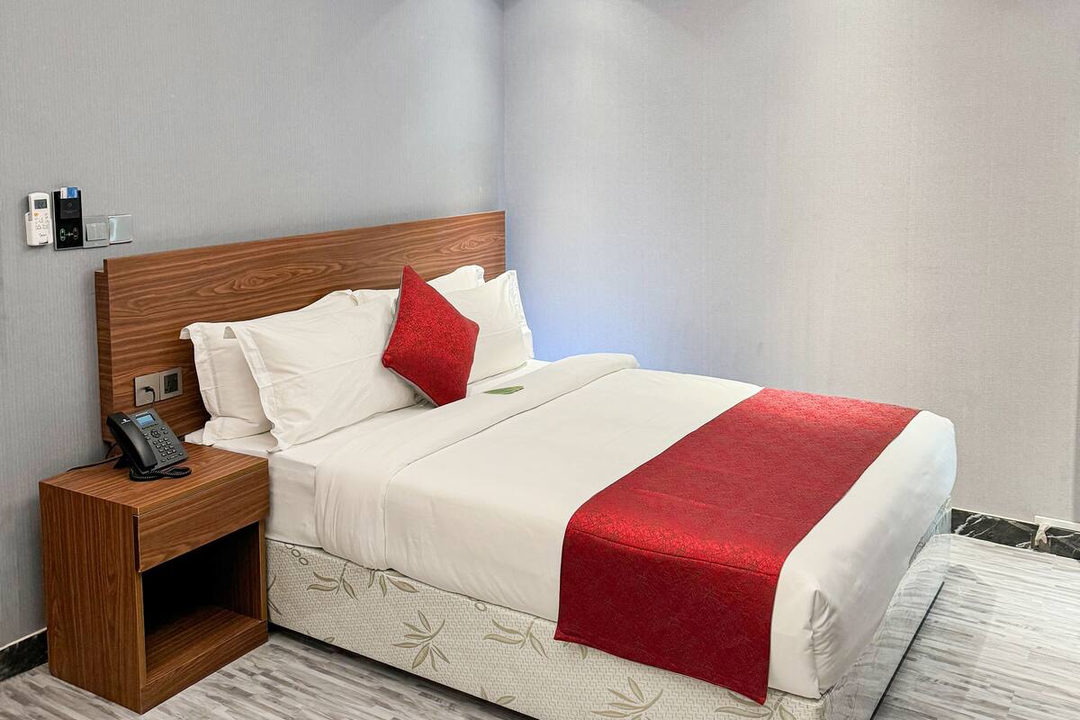 Una habitación de hotel con una cama bien hecha, una mesita de noche de madera con un teléfono y un termostato fijado a la pared.