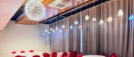Un salón de banquetes con mesas redondas cubiertas con manteles blancos y fundas rojas para las sillas, con un telón de fondo de cortinas grises y un techo de madera con una lámpara de diseño geométrico.