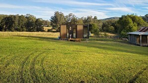A modern, single-story cabin with a wooden exterior, situated in a grassy field with trees in the background. - Tiny Ironbark by Tiny Away (Glenreagh)