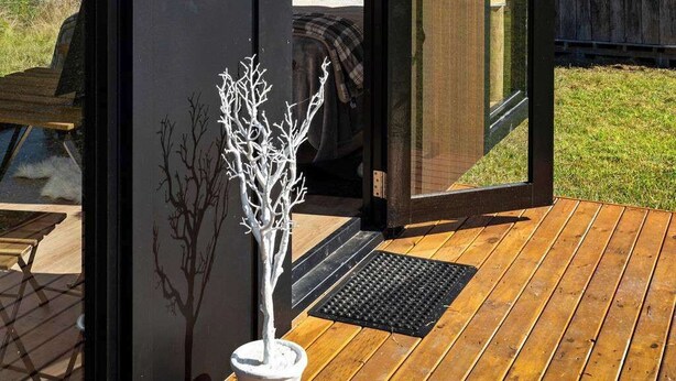 A modern, compact cabin with a sliding glass door leading to a bedroom with a bed and a nightstand. - Tiny Ironbark by Tiny Away (Glenreagh)