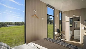 A modern bedroom with a large window, a bed, a desk, and a view of a grassy field. - Tiny Ironbark by Tiny Away (Glenreagh)