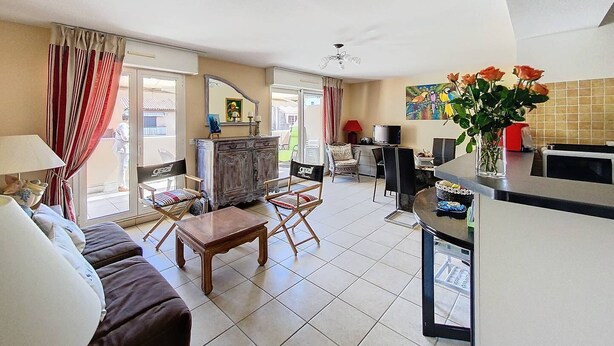 A spacious living area with a sofa, armchairs, a coffee table, and a dining table. There is a kitchen area with a refrigerator, microwave, and a vase with flowers.