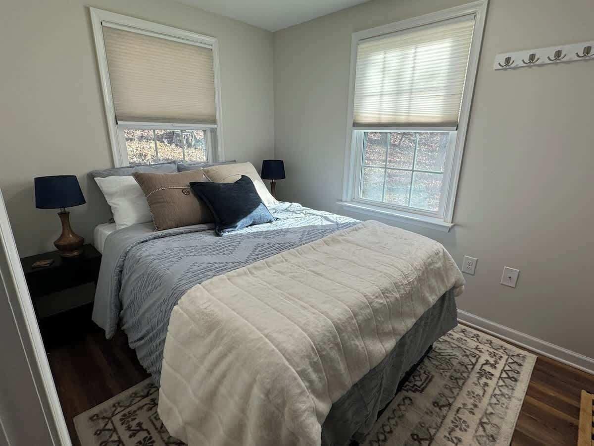 Un dormitorio con una cama, dos mesitas de noche, una mesita auxiliar, una ventana con persianas y una alfombra estampada.