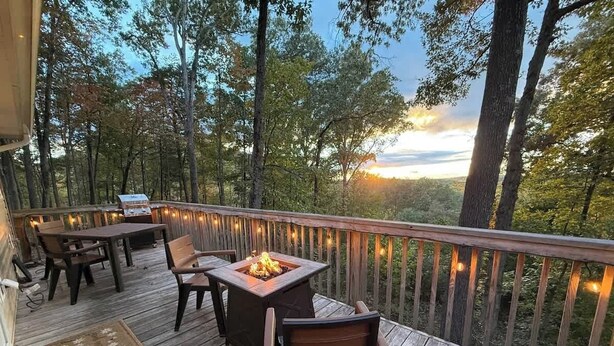 Una terraza de madera con mobiliario exterior y un brasero, rodeada de árboles y una barandilla.