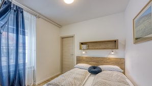 Une chambre avec une tête de lit en bois, une table de chevet, un lit avec une literie à motifs, une fenêtre avec des rideaux légers et une étagère murale remplie de livres.