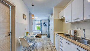 Een smalle gang die leidt naar een slaapkamer met een bed, een kleine tafel met stoelen, een raam met gordijnen, en een kitchenette met een spoelbak en kastjes.