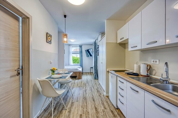Un couloir étroit mène à une chambre comprenant un lit, une petite table avec des chaises, une fenêtre avec des rideaux et une kitchenette équipée d’un évier et de placards.