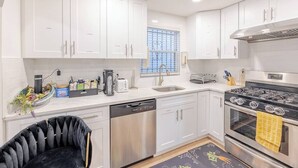 A modern kitchen with white cabinets, a stainless steel dishwasher, a gas stove, and a window with blue blinds. - Exquisite 3 Bedrooms (Newark)