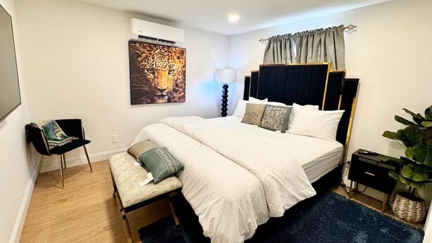 A modern bedroom with a large bed, bedside tables, a chair, a potted plant, and a wall-mounted air conditioner. - Exquisite 3 Bedrooms (Newark)