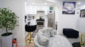 A modern kitchen with white cabinets, a microwave, and a refrigerator. There is a dining area with a patterned sofa and a black ottoman. A large painting is on the wall, and a potted plant is in the corner. - Exquisite 3 Bedrooms (Newark)
