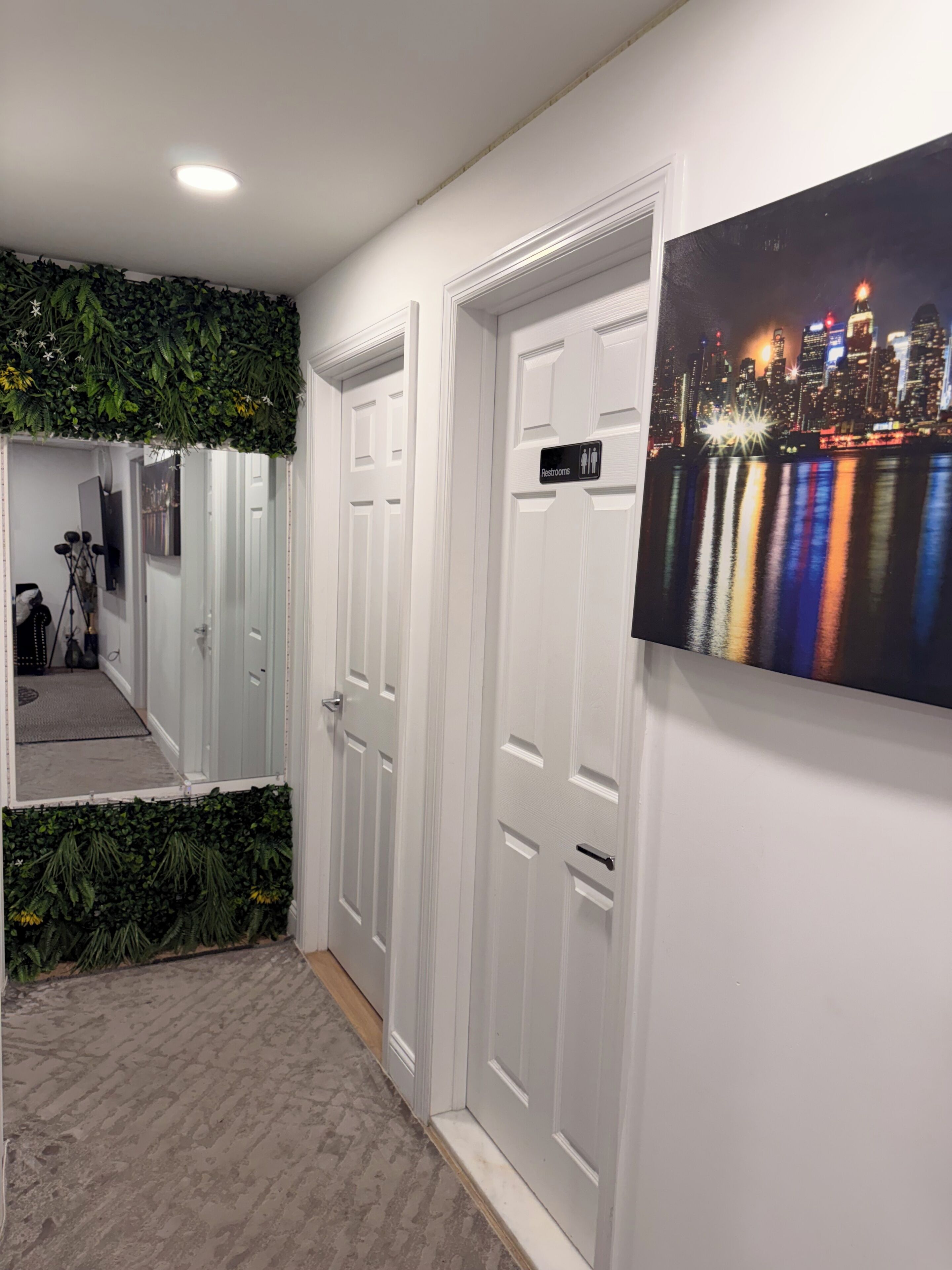 A hallway with white doors, a cityscape mural, and a green plant wall.