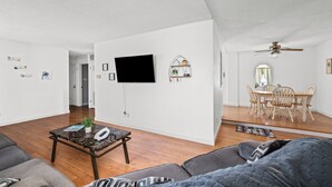 A spacious living room with a flat-screen TV, a dining area with a wooden table and chairs, and a ceiling fan.