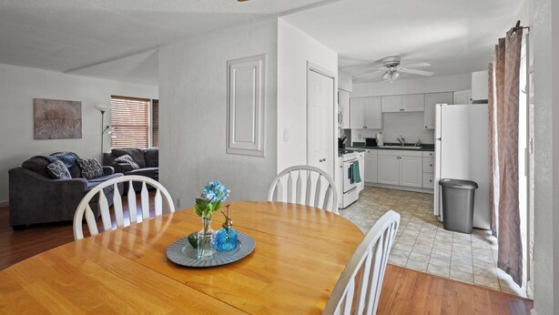 A dining area with a wooden table, white chairs, and a vase with flowers. There is a kitchen in the background with white cabinets and a refrigerator.