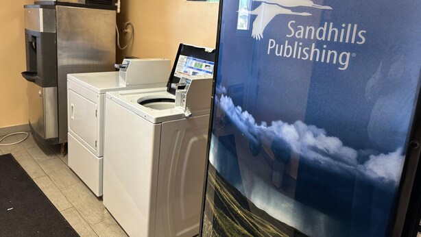 A room with a large screen displaying Sandhills Publishing, a commercial washer and dryer unit, and a wall-mounted air conditioning unit. - Stay Inn by OYO Near I 80 Lincoln West (Lincoln)