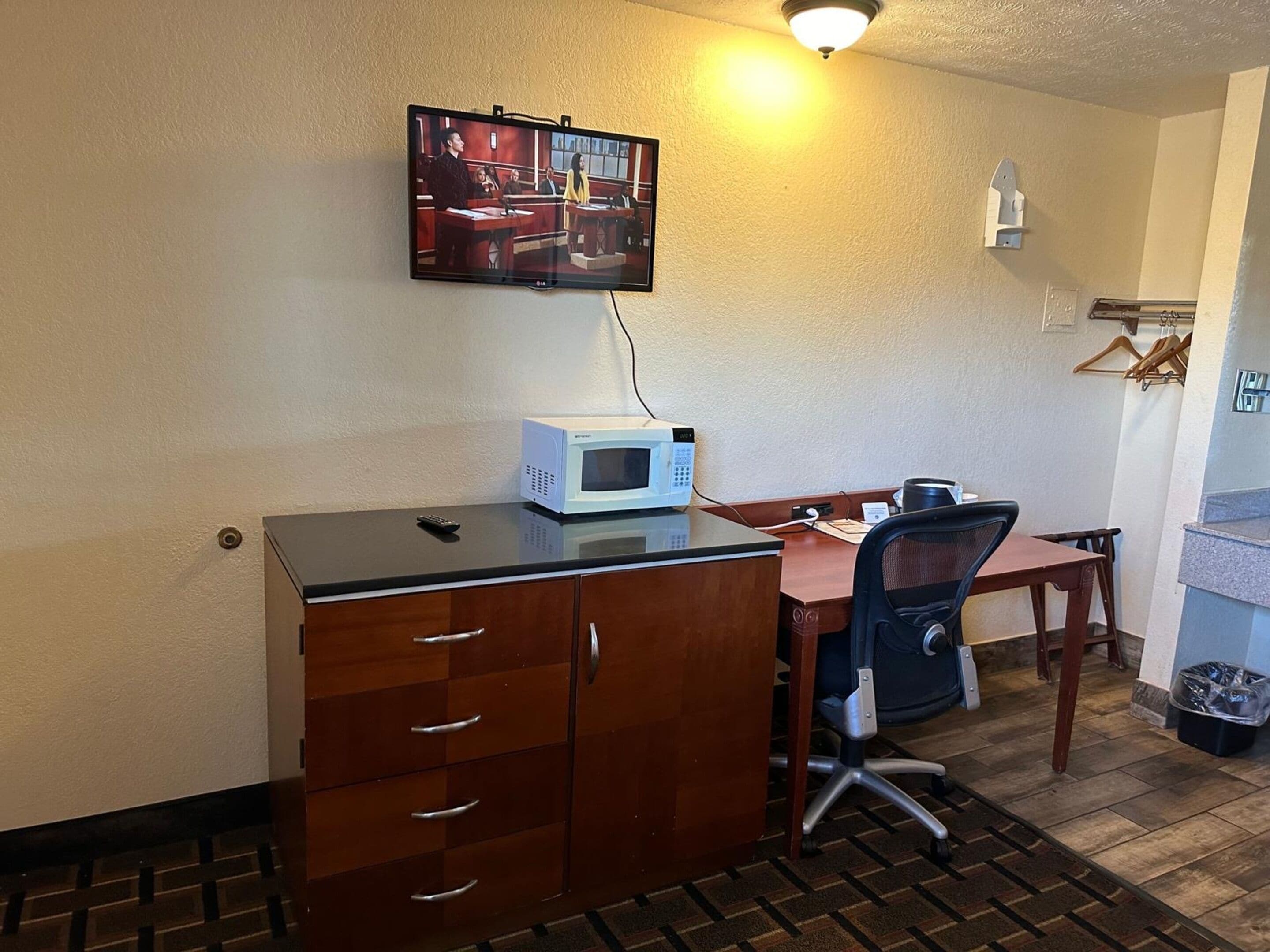 A compact hotel room with a kitchenette, a desk with a chair, a television mounted on the wall, and a microwave on the counter.