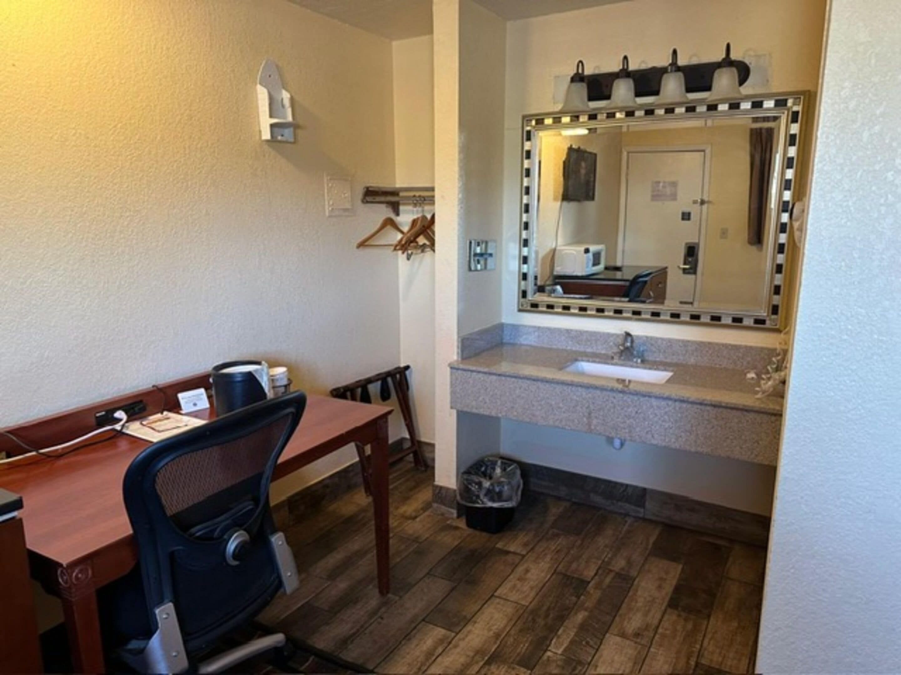 A hotel room with a wooden desk, a black office chair, a small trash bin, a sink with a mirror above it, and a light fixture on the wall.