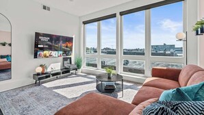 A modern living room with a large TV, comfortable seating, a coffee table, and a view of the city through large windows. - FIFA 10 Mins Away | Rooftop | Gym | King bed (Philadelphia)