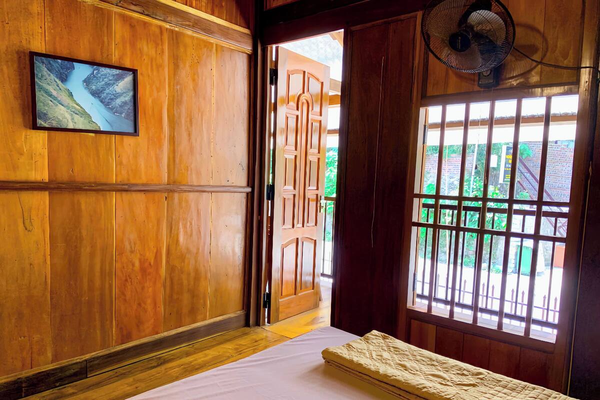 A wooden room with a bed, a fan, a window with bars, and a framed picture on the wall.