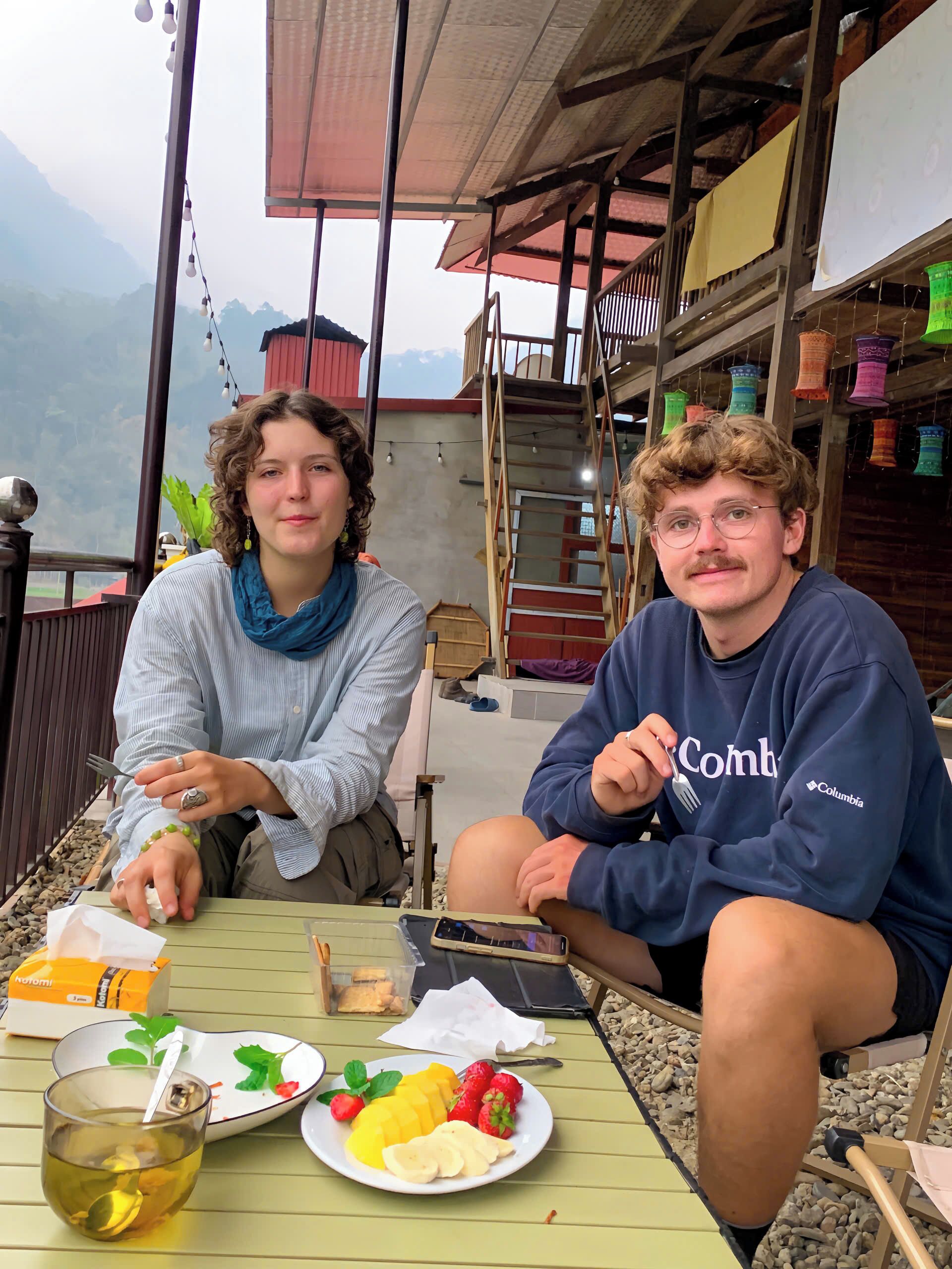 Zwei Personen sitzen an einem Tisch im Freien mit einer Fruchtschale und einer Teetasse.