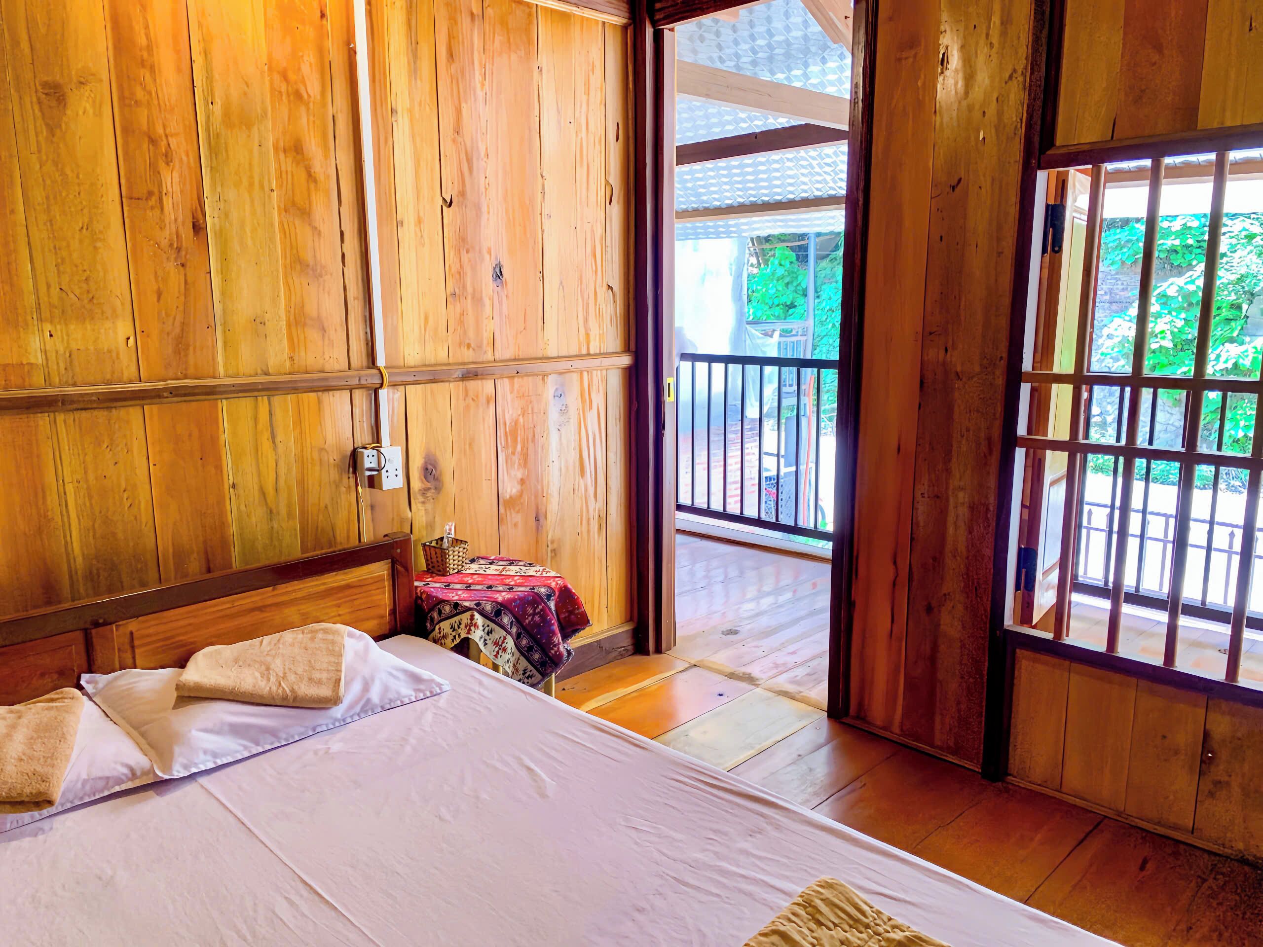 Une pièce en bois avec un lit, une petite table recouverte d’une nappe à motifs et une vue sur la verdure à l’extérieur.