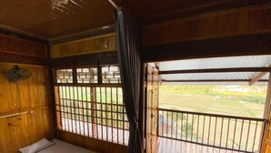 A room with a wooden interior, a bed, a window with a view of a green landscape, and a balcony with a metal railing. - Duy Tuyen Ba Be Homestay Free BBQ (Ba Be)