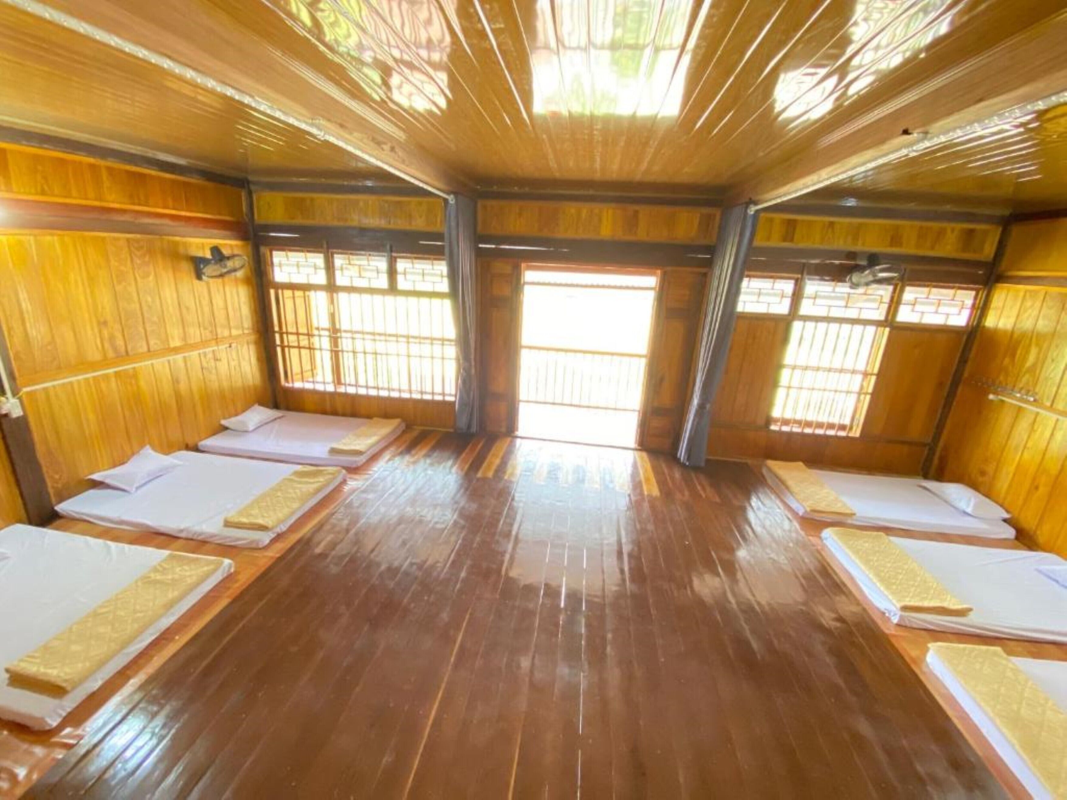 A room with wooden floors and walls, multiple beds with white linens, and large windows allowing natural light.