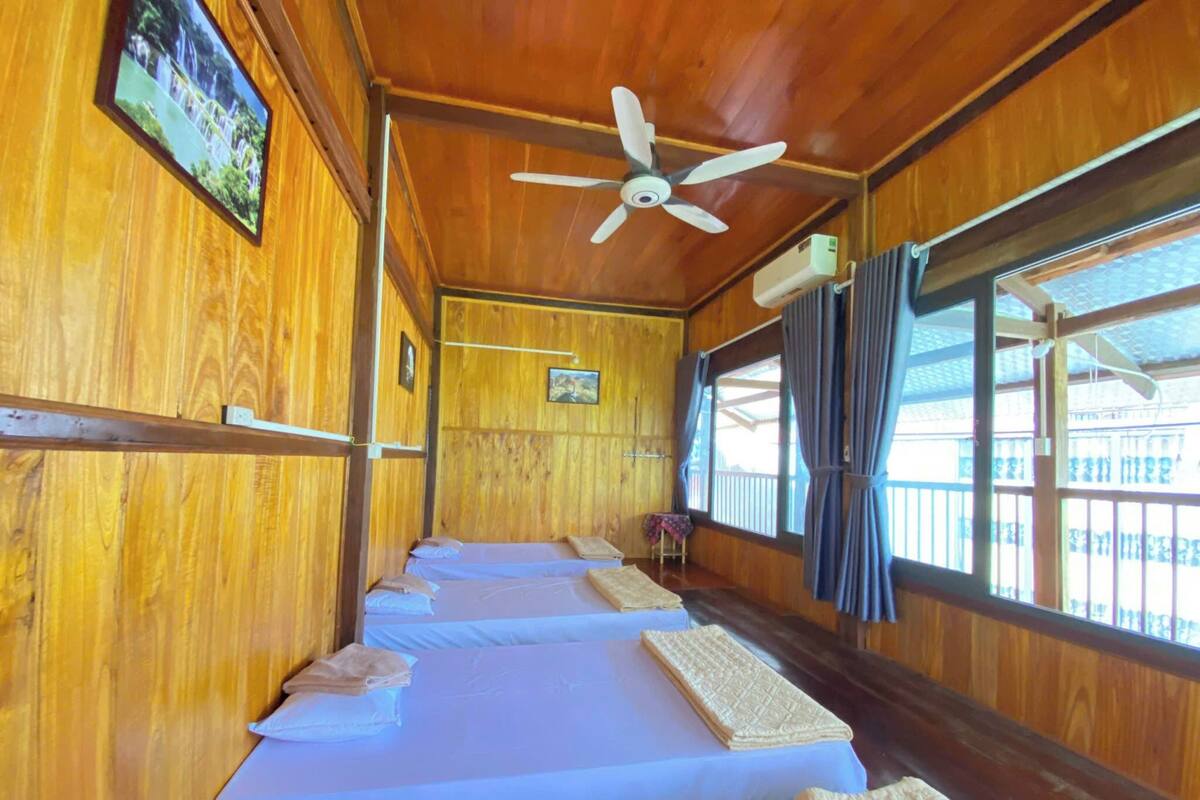 A row of wooden bunk beds with a ceiling fan, a small table with a chair, and a window with curtains.