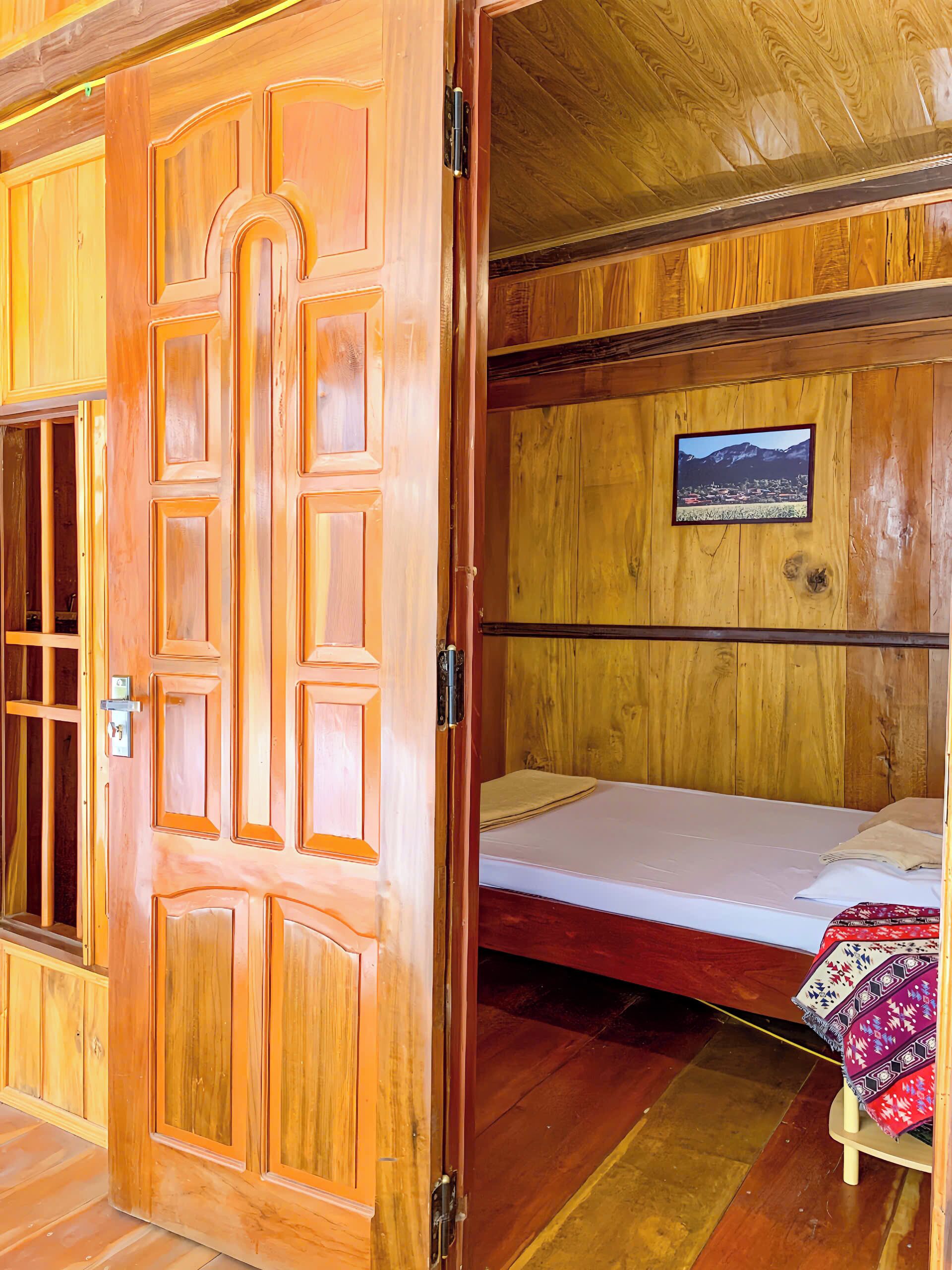 A wooden room with a bed, a small table with a cloth, and a picture on the wall.