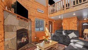 Un acogedor interior de cabaña con chimenea de piedra, un sofá gris suave, un banco de madera y vistas del paisaje nevado.