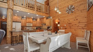 Un interior rústico de madera con un comedor que incluye una mesa blanca y bancos, una cocina con armarios de madera, y un altillo con barandilla de madera.