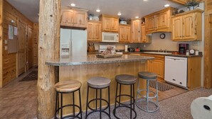 Una cocina con armarios de madera, encimera de granito y un refrigerador. Hay tres taburetes y un microondas.