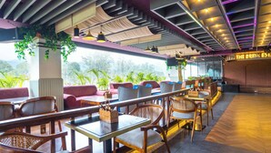 A restaurant with a modern interior, featuring wooden tables and chairs, a brick wall with a neon sign, and large windows overlooking a green landscape. - Treebo White Sand (Dehradun)