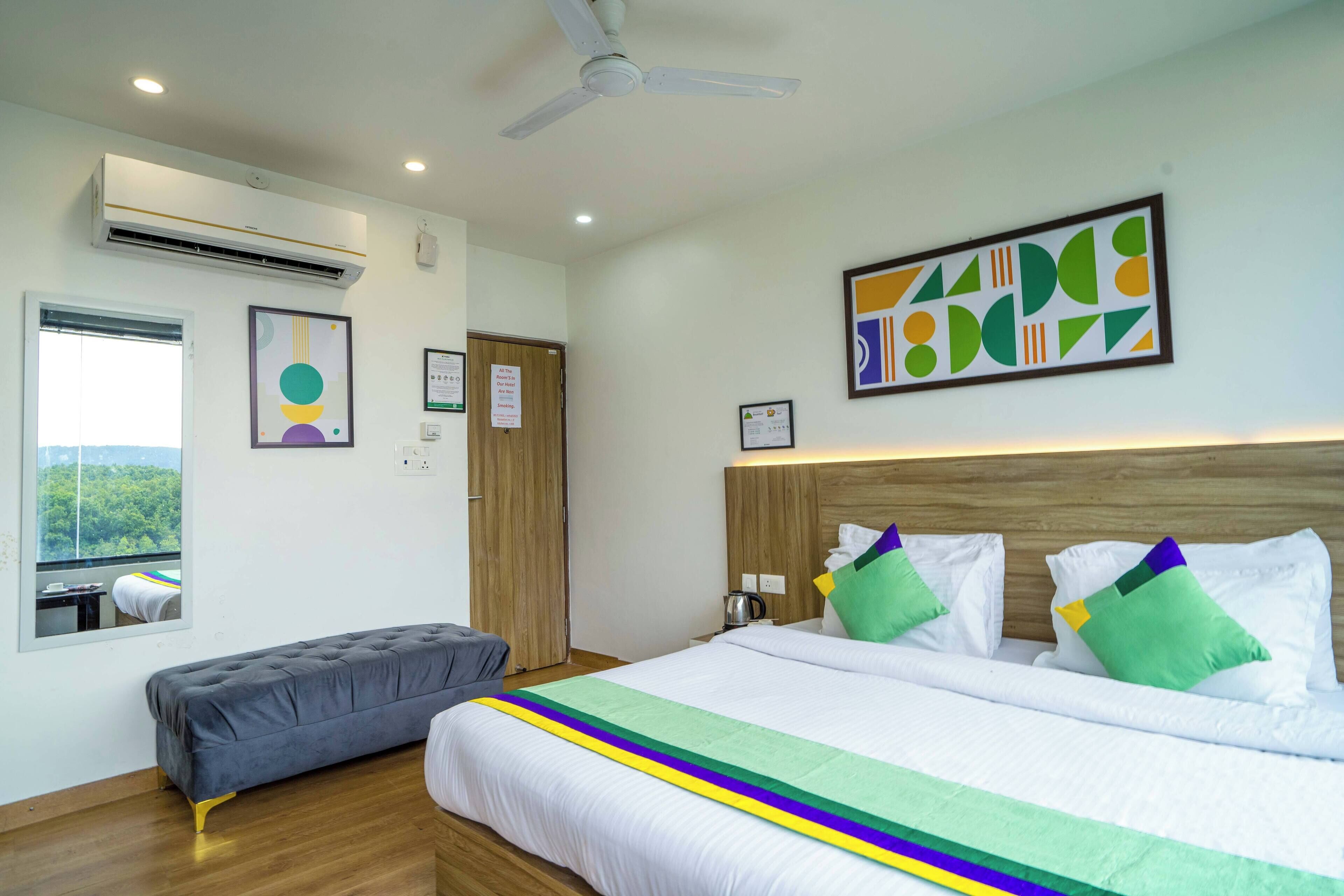 Habitación de hotel con una cama grande, una cama más pequeña, un banco, equipo de aire acondicionado, ventilador de techo y ventana con vista a la vegetación.