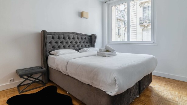 A bedroom with a tufted headboard, a neatly made bed, a small stool, and a window with a view of buildings. - 3 pieces Arc de triomphe Champs Elysée - Paris 16 (Paris)
