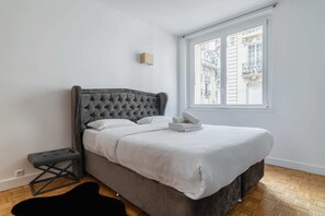 Une chambre à coucher avec une tête de lit capitonnée, un lit bien fait, un petit tabouret et une fenêtre donnant sur des immeubles.