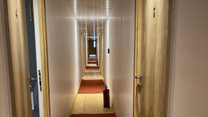 Un largo y estrecho pasillo de hotel con puertas de madera a ambos lados, una alfombra roja y un extintor en el centro.