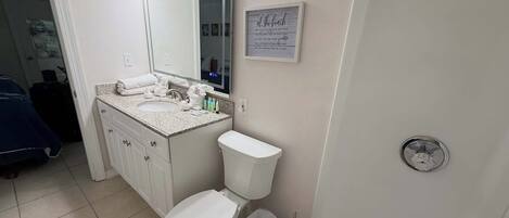 Casa de banho com lavatório branco, espelho e sanita. Há um contentor de lixo e um porta-toalhas.