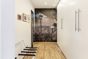 A hallway with a wooden floor, white walls, and a large floral mural. There are framed pictures on the wall, a white cabinet with silver handles, and a metal bench.