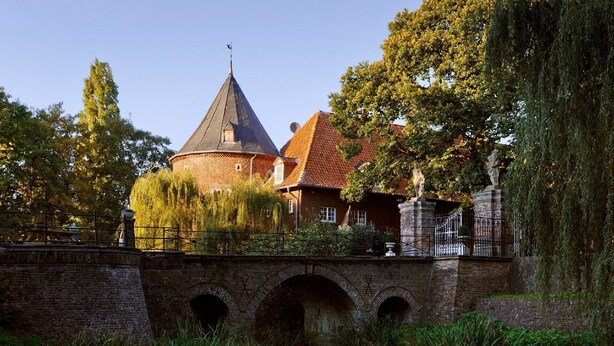 Ein historisches Gebäude mit einem markanten Turm, einer Ziegelbrücke und üppigem Grün.