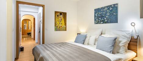 A hotel room with a bed, a nightstand, a painting, and a hallway leading to another room.