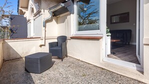 Ein überdachter Außenbereich mit Sessel und Hocker, einem Fenster mit Jalousien und einer Tür zu einem Innenraum.