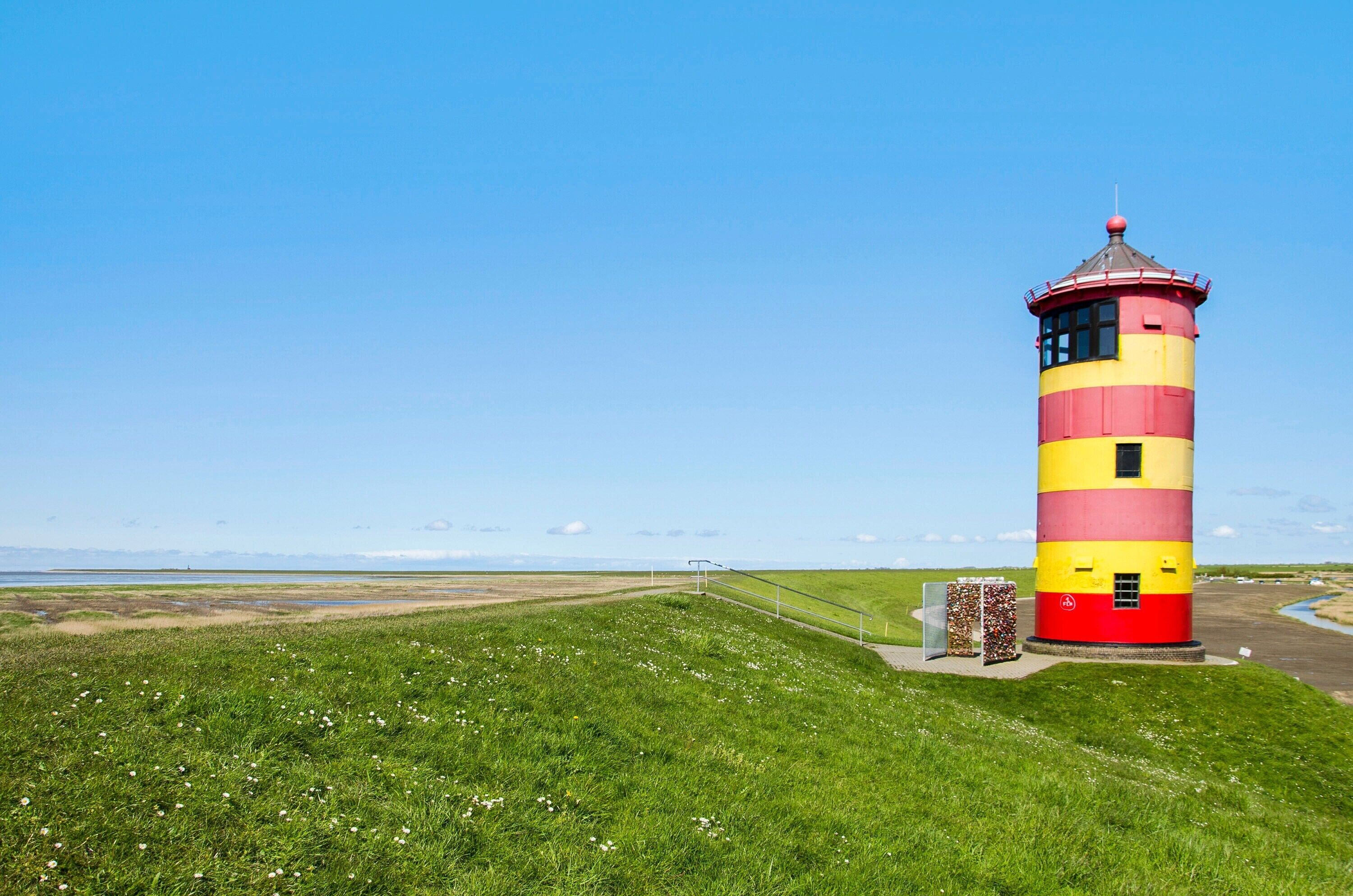 A lighthouse with a red and yellow striped exterior, situated on a grassy hill with a clear sky.