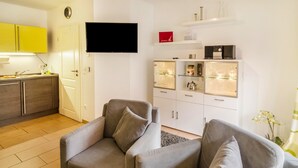 A living room with two grey sofas, a flat-screen TV, a white cabinet with glass doors, and a kitchen area with yellow cabinets.